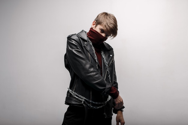 A young man with a metal chain with a fashionable hairstyle in a red stylish golf in a black leather stylish jacket in black stylish jeans poses indoors against a white wall. Stylish handsome guy