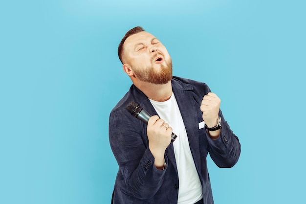 Young man with microphone on blue studio background, leading concept. Human emotions and facial expressions concepts.