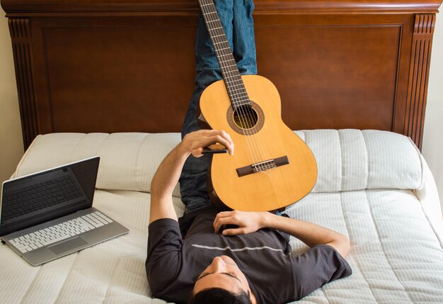 Photo young man with mobile phone and guitar resting on bed