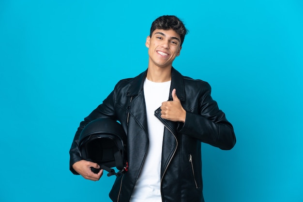 Young man with a motorcycle helmet over blue giving a thumbs up gesture