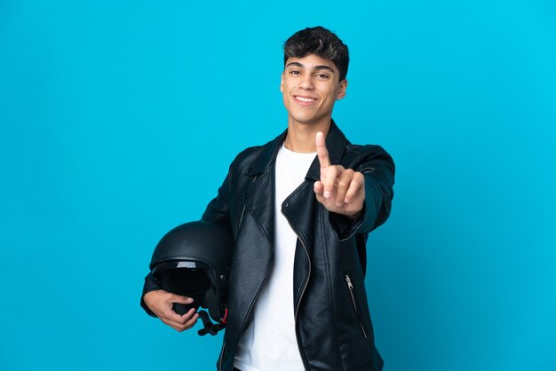 Young man with a motorcycle helmet over isolated blue background showing and lifting a finger