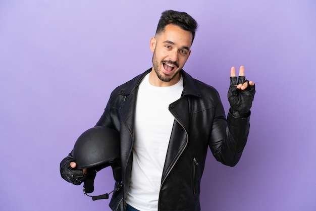 Young man with a motorcycle helmet isolated on purple background smiling and showing victory sign