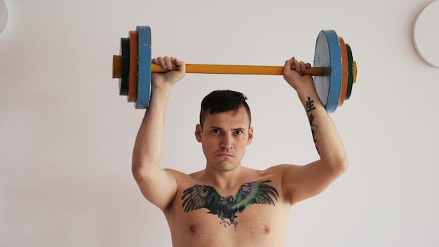 Young man with naked body and tattoo on chest lifting multicolored barbell against white patterned wall Adult strong guy doing sport at home