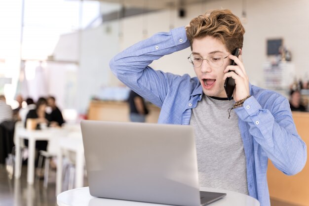 Young man with piercings talking on a mobile phone and with a surprised expression sitting at a table with a laptop working on a coworking