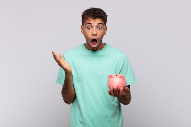 Young man with a piggy bank feeling extremely shocked and surprised, anxious and panicking, with a stressed and horrified look