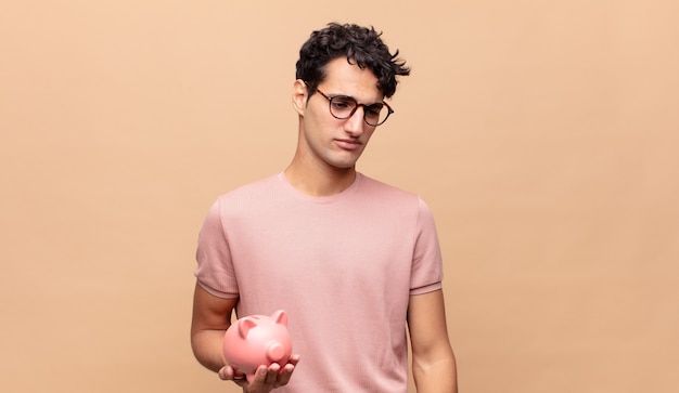 Young man with a piggy bank feeling sad, upset or angry and looking to the side with a negative attitude, frowning in disagreement