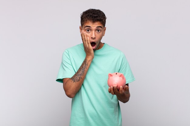 Young man with a piggy bank feeling shocked and scared, looking terrified with open mouth and hands on cheeks