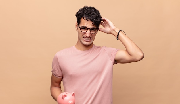 Young man with a piggy bank feeling stressed, worried, anxious or scared, with hands on head, panicking at mistake