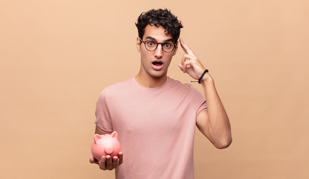 Young man with a piggy bank looking surprised, open-mouthed, shocked, realizing a new thought, idea or concept