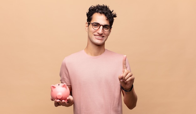 Young man with a piggy bank smiling proudly and confidently making number one pose triumphantly, feeling like a leader