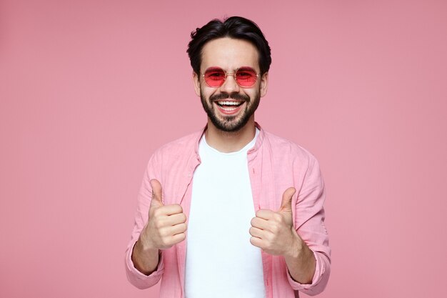 Young man with pink sunglasses smiling and showing thumb up gesture with both hands while standing on pink background