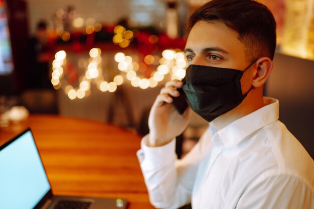 Young man with protective face mask working on laptop at home during winter holiday.