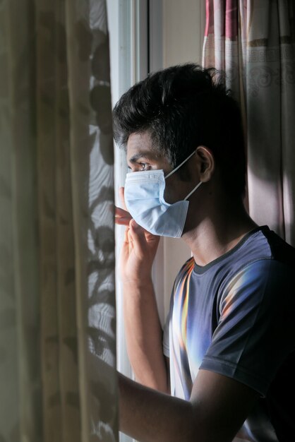 Молодой человек в защитной маске смотрит в окно.