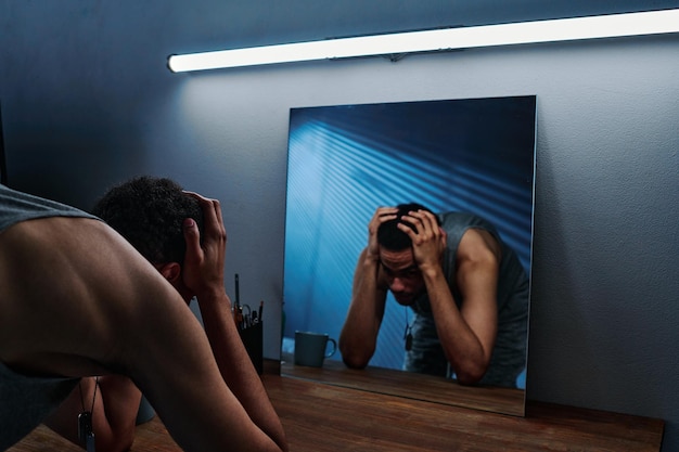 Young man with ptsd standing in front of mirror with his head in hands