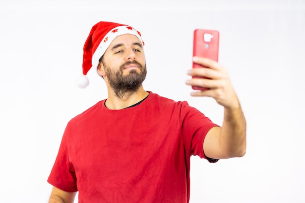 Young man with the red christmas hat taking a selfie on a white background, dressed in a red t-shirt, copy paste