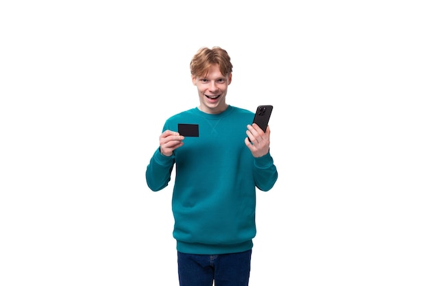 A young man with red hair dressed in a blue sweater holds a credit card and a smartphone and smiles