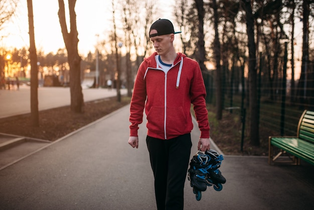 Young man with roller skates in hands walking in city park. Male rollerskater leisure