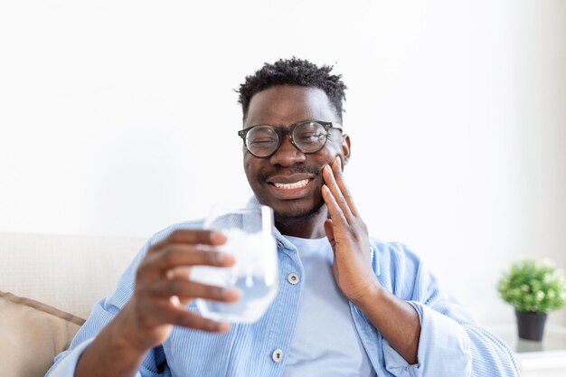 Young man with sensitive teeth and hand holding glass of cold water with ice Healthcare concept man drinking cold drink glass full of ice cubes and feels toothache pain