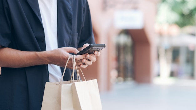 買い物袋を持つ若い男は買い物をしながら携帯電話を使用しています。