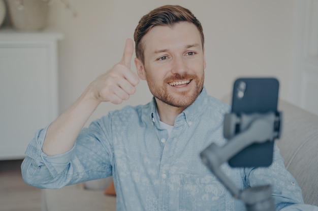 Young man with short beard showing thumbs up gesture while video chatting on phone attached to gimbal indoors, talking with his relatives, telling them that everything is fine