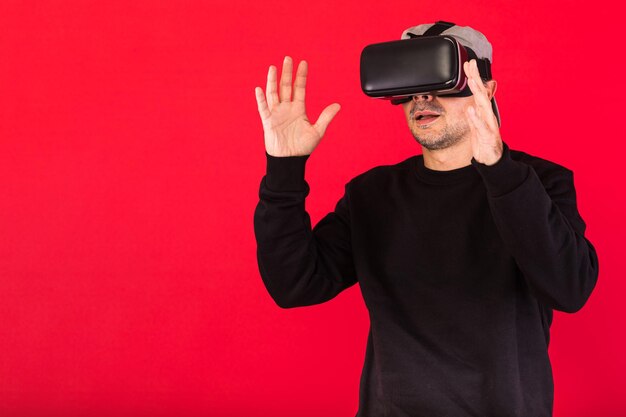 Young man with short beard, wearing black sweatshirt, cap and\
virtual reality glasses amazed, touching something virtually, on\
red background. technology, vr, computing and hobbies concept.