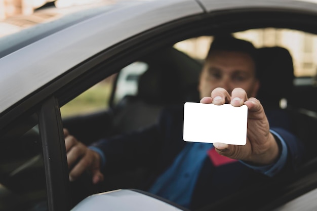 Young man with sitting in car showing an empty white card. Driver has got driving license. Parking pass or entrance.