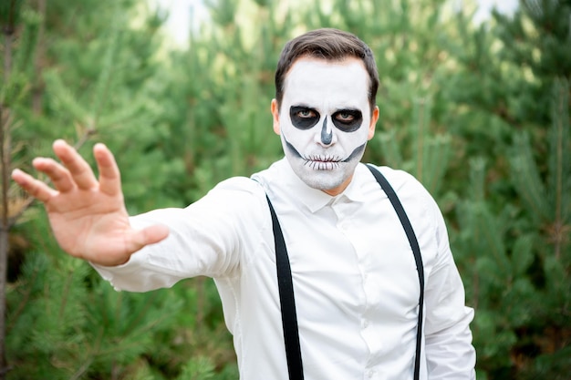 Молодой человек со скелетным макияжем стоит в лесу на вечеринке в честь Хэллоуина