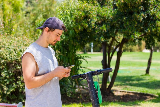 晴れた日に公園でスクーターでスマートフォンを持つ若い男代替通勤輸送の概念