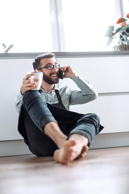 Young man with smartphone sitting on kitchen floor