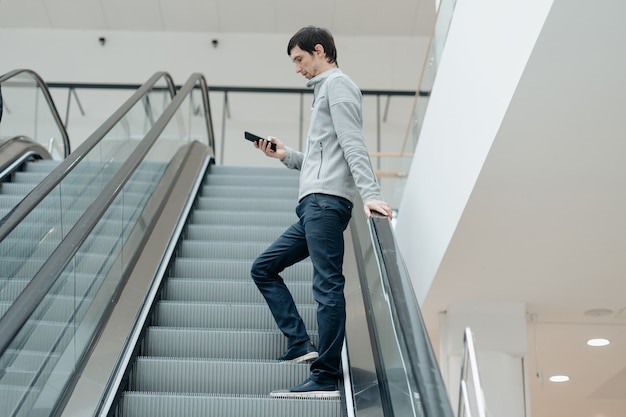 エスカレーターの近くに立っている誰かを待っているスマートフォンを持つ若い男