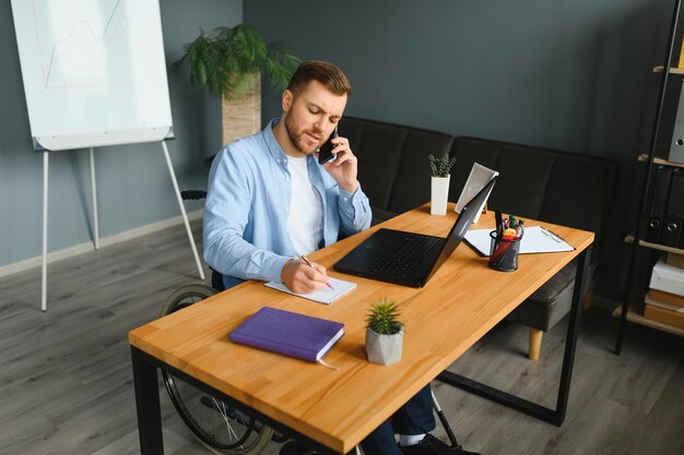 Young man with special needs in casual clothes working on wireless laptop Male freelancer working from home while sitting in wheelchair