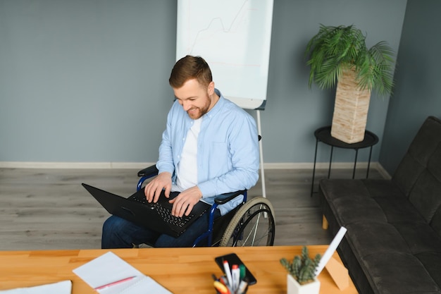 Foto giovane con bisogni speciali in abiti casual che lavora su laptop wireless libero professionista maschio che lavora da casa mentre è seduto su una sedia a rotelle