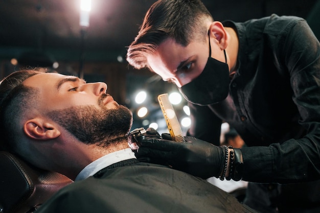 Young man with stylish hairstyle sitting and getting his beard shaved by guy in black protective mask in barber shop