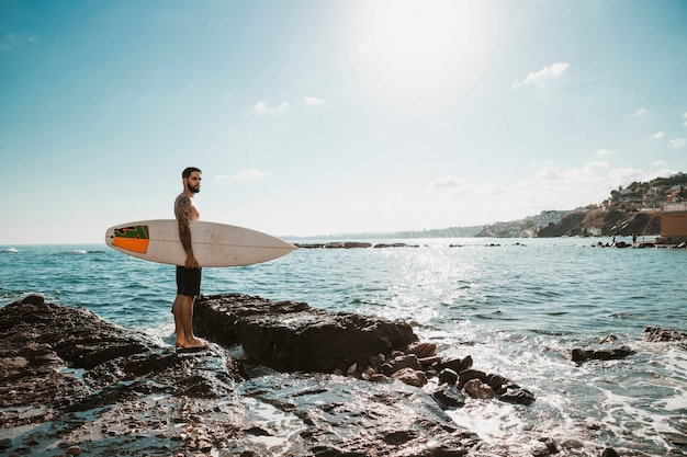 Young man with surf board on stone near water