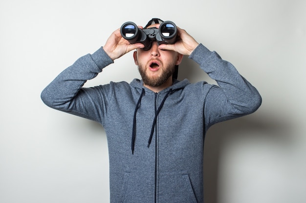 Young man with a surprised face in a hoodie and a cap looks through binoculars against a light wall