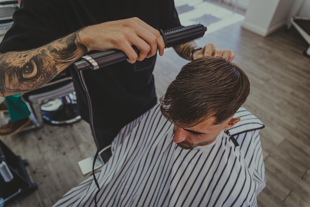 A young man with tattooed arms cuts a man's hair in a barbershop