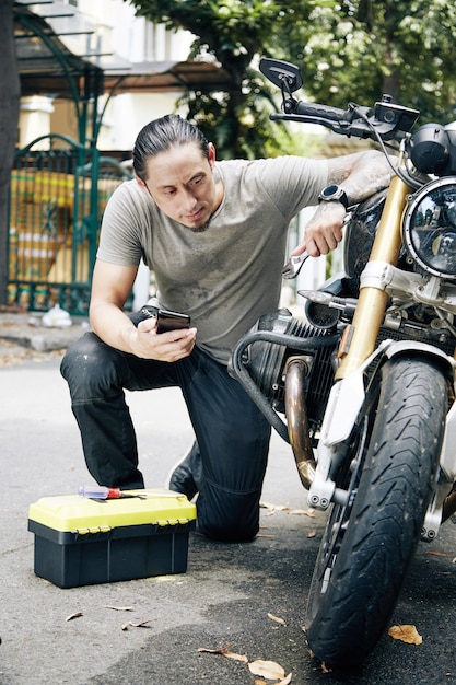 Young man with toolbox following tutorial on smartphone when repairing his broken motorcycle