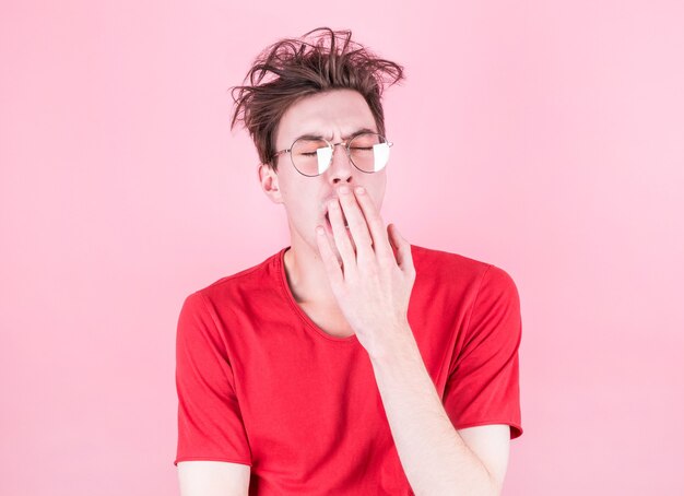 Young man with tousled hair yawns from fatigue against a pink wall. Stress, fatigue and sleep concept.