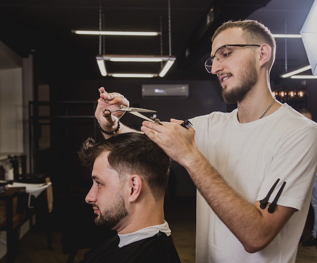 Young man with trendy haircut at barber shop. Barber does the hairstyle and beard trim.