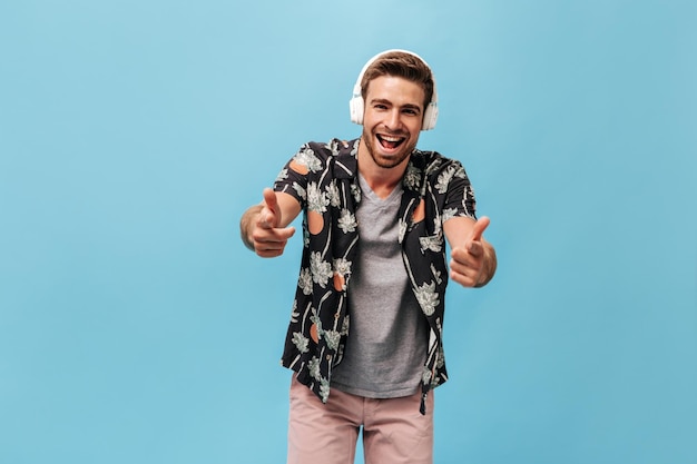 Young man with trendy hairstyle in grey tshirt printed shirt and light shorts looking into camera with smile and listening to music