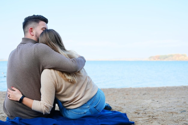 A young man and woman are sitting against the backdrop of a\
large blue lake and hugging couple sitting with their backs to the\
camera