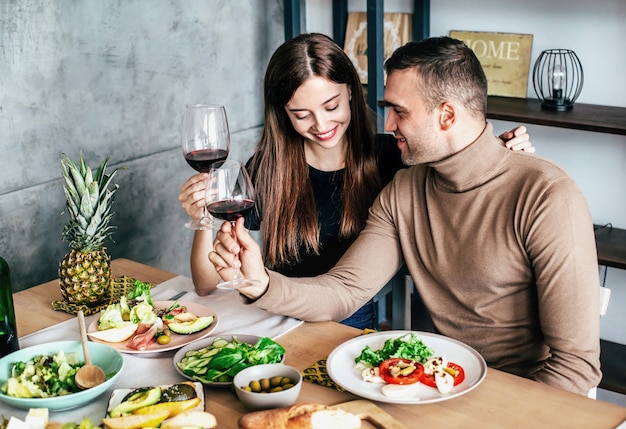 Young man and woman are sitting at a table covered with food and drinks with glasses of wine in their hands