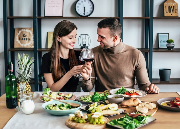 young man and woman are sitting at a table covered with food and drinks with glasses of wine in their hands