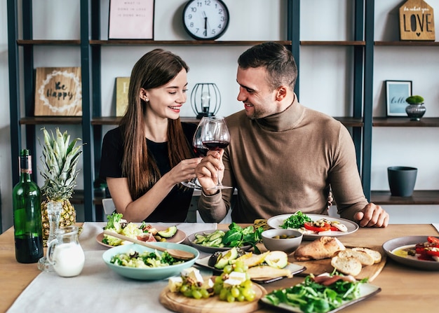 Young man and woman are sitting at a table covered with food and drinks with glasses of wine in their hands