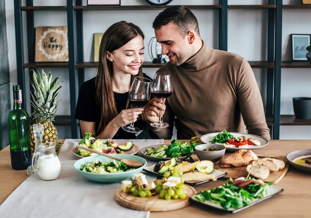 Young man and woman are sitting at a table covered with food and drinks with glasses of wine in their hands