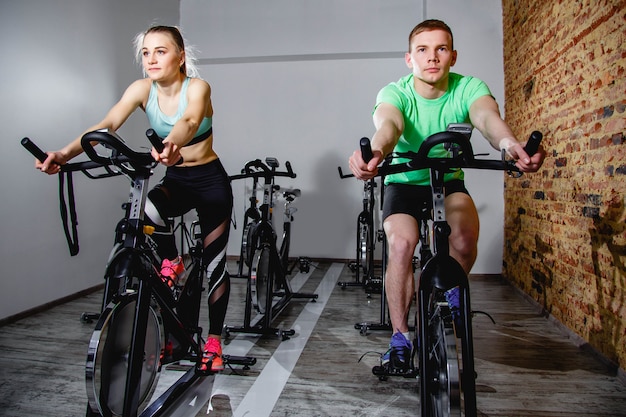 Young man and woman biking in the gym, exercising legs doing cardio workout cycling bikes.