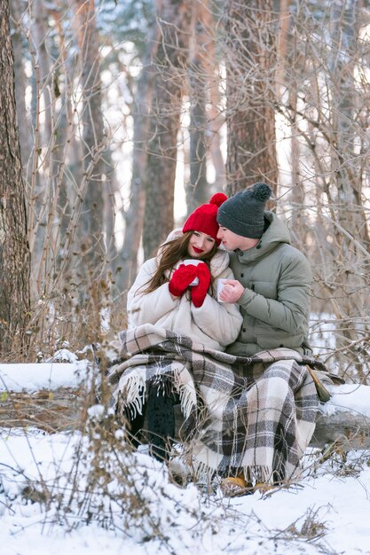 Young man and woman had picnic in the park in winter. Lovers under warm blanket drink tea in snowy forest. Vertical frame