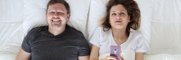 Photo young man and woman lying in bed and holding phone and joystick top view family leisure concept