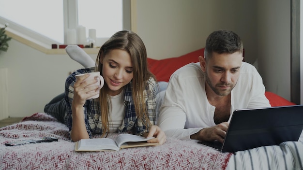 젊은 남자와 여자는 침대에 누워 책을 읽고 아침에 노트북 컴퓨터를 사용합니다