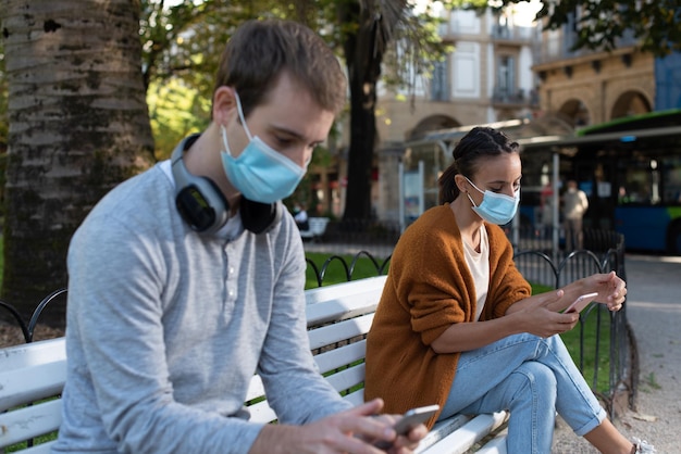 Young man and woman sitting on a bench checking their phones while keeping social distance during Coronavirus pandemic Couple wearing face masks